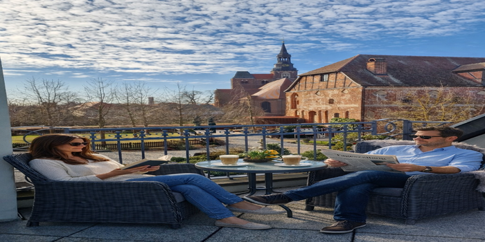 Paar, lesend auf der Clubzimmerterrasse sitzend. Zwischen Ihnen ein Tisch mit Kaffee und im Hintergrund die St. Stephanskirche. Teilweise KI-modifiziert: Die dargestellten Personen wurden mithilfe generativer KI in eine Originalaufnahme unseres Hotels beziehungsweise der Umgebung eingefügt.